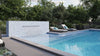 Modern outdoor pool area with a white cascading water wall, lounge chairs, and lush greenery in the background, enhanced by the subtle beauty of mosaic tiles.