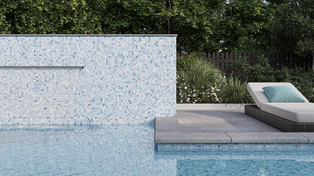 Poolside view with a stunning mosaic tile wall, featuring a waterfall and a lounge chair adorned with a turquoise pillow. Lush greenery completes the tranquil setting.